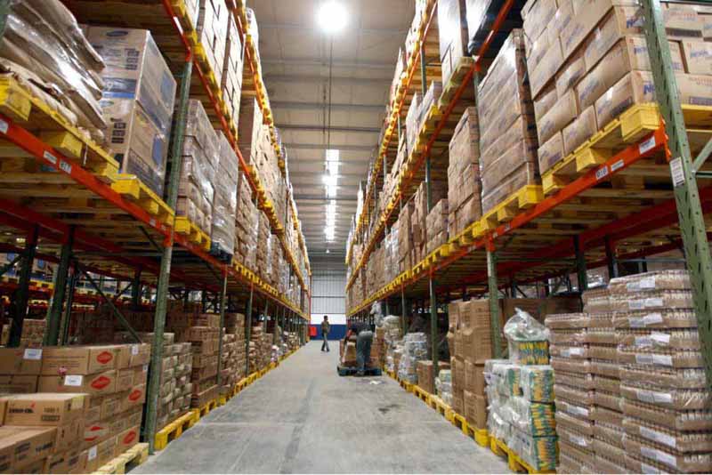 Tall shelves stacked with boxes stand in a spacious, well-lit warehouse as two workers move goods using a cart.