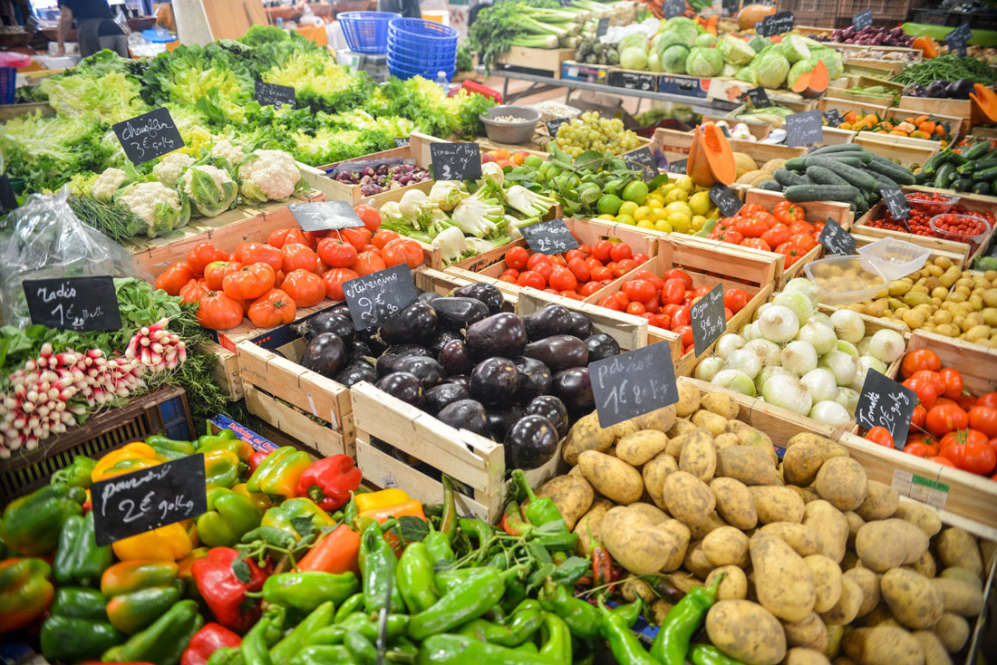 Fresh fruits and vegetables displayed in wooden crates at a farmers' market. Chalkboard signs list names and prices.