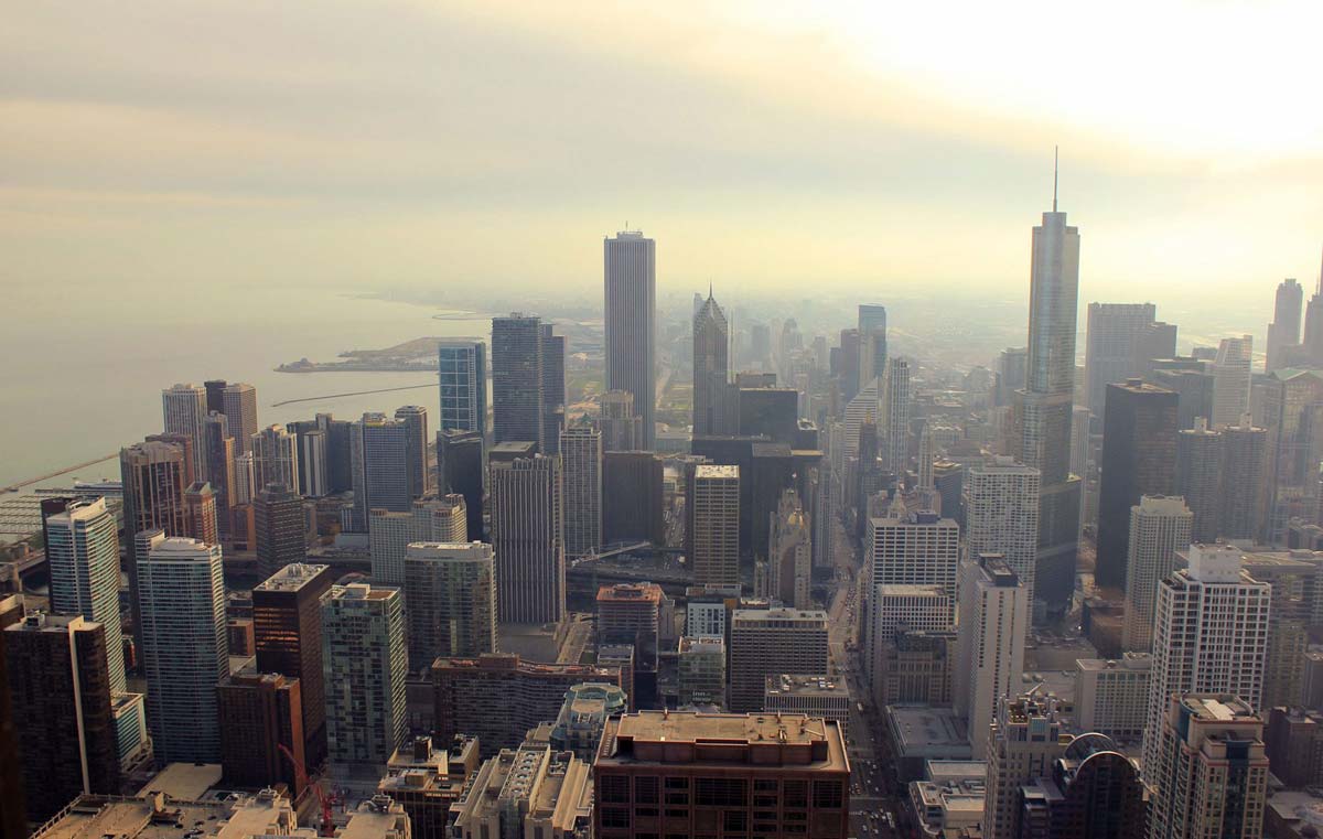 Skyscrapers standing tall amidst an urban landscape, with hazy skies overhead and a large body of water in the background.