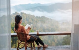 A woman sits in a wicker chair reading a book with a mug while overlooking a countryside horizon.