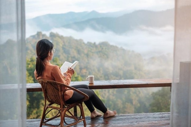 A woman sits in a wicker chair reading a book with a mug while overlooking a countryside horizon.